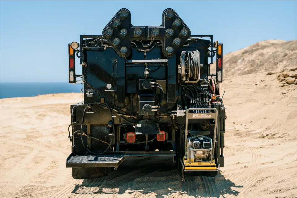 Rear view of the patcher truck showing the light arrow board, the hose reel and a small plate compactor on the step, with the ocean on the horizon