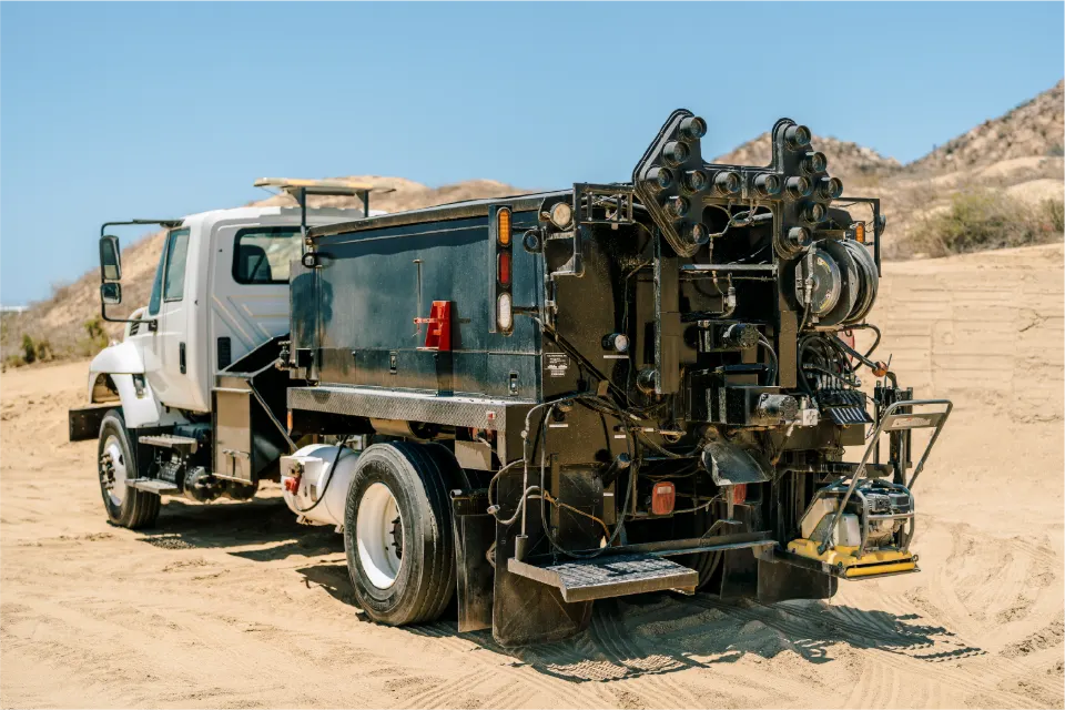 The pothole patcher viewed from its rear corner, the black body lined with hoses, burners and an arrow board, standing on sand beneath dry desert hills
