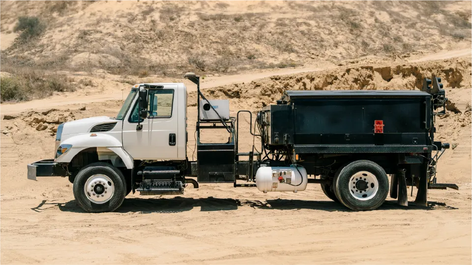Side view of the white International truck with its Pro Patch 4000 unit and a propane tank, parked on a graded dirt lot under a sandy bank
