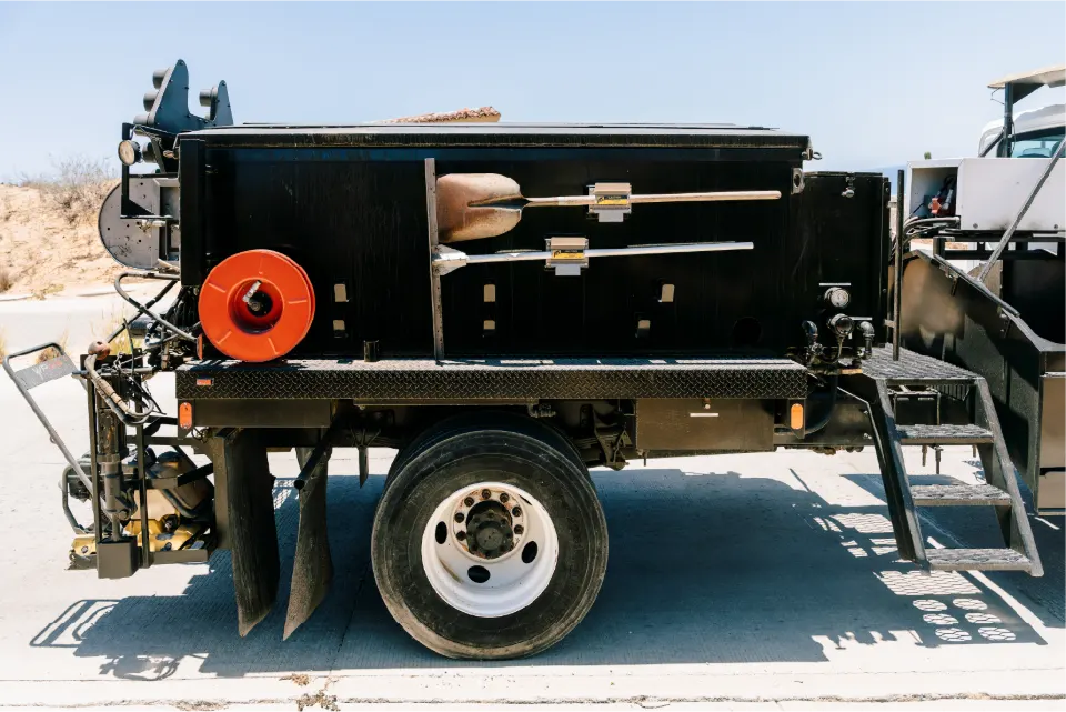 Close side view of the pothole patcher body showing shovels clipped to the side, a red hose reel, mud flaps and a folding step