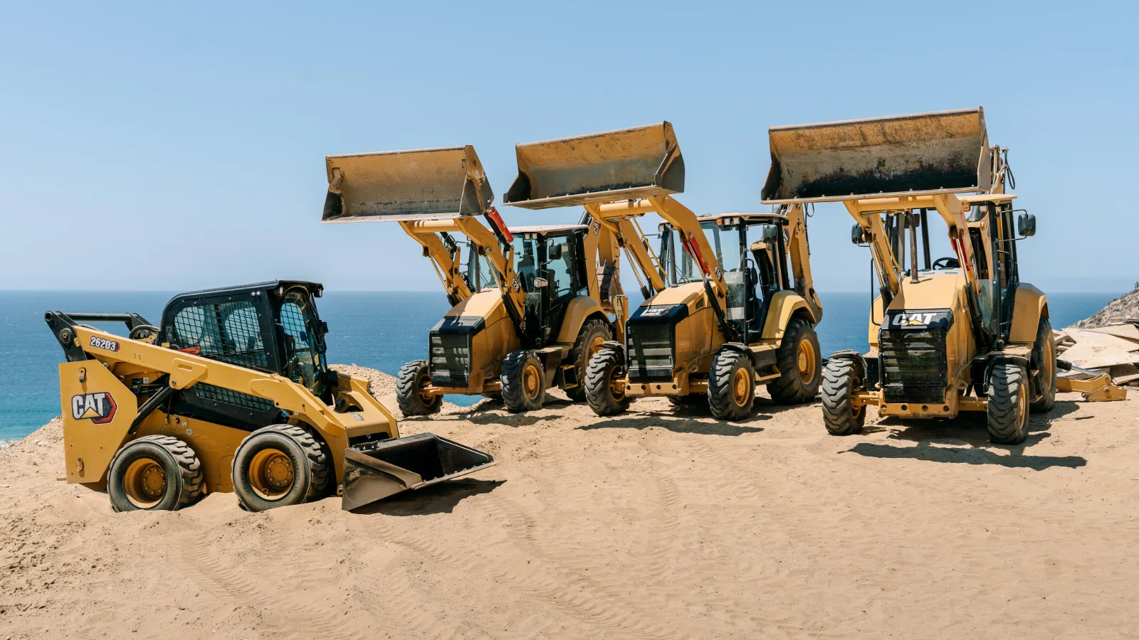 Creatio Construction fleet of CAT backhoe loaders and a skid steer lined up on a beach in Los Cabos