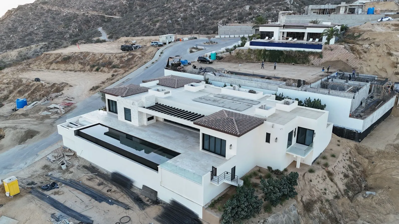 Aerial view of a large white home under construction on a desert hillside, with more homes on the slope above