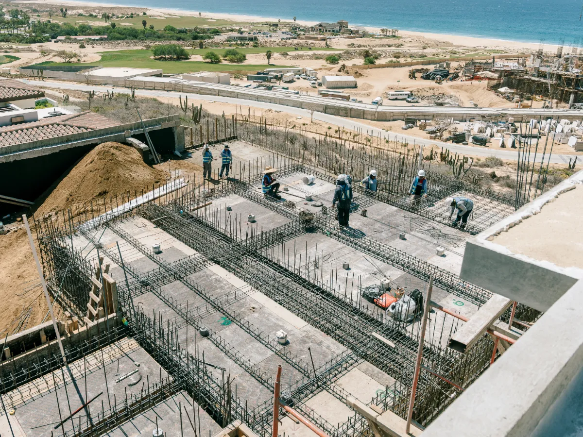 Workers tying steel reinforcement for a concrete foundation on a coastal building site, with the beach in the background
