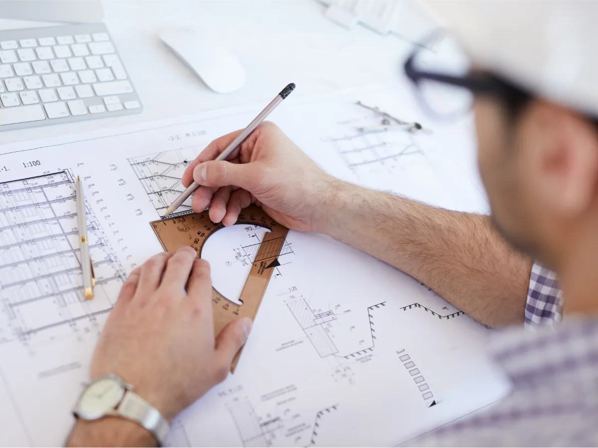 Engineer in a white hard hat measuring technical drawings with a protractor and pencil