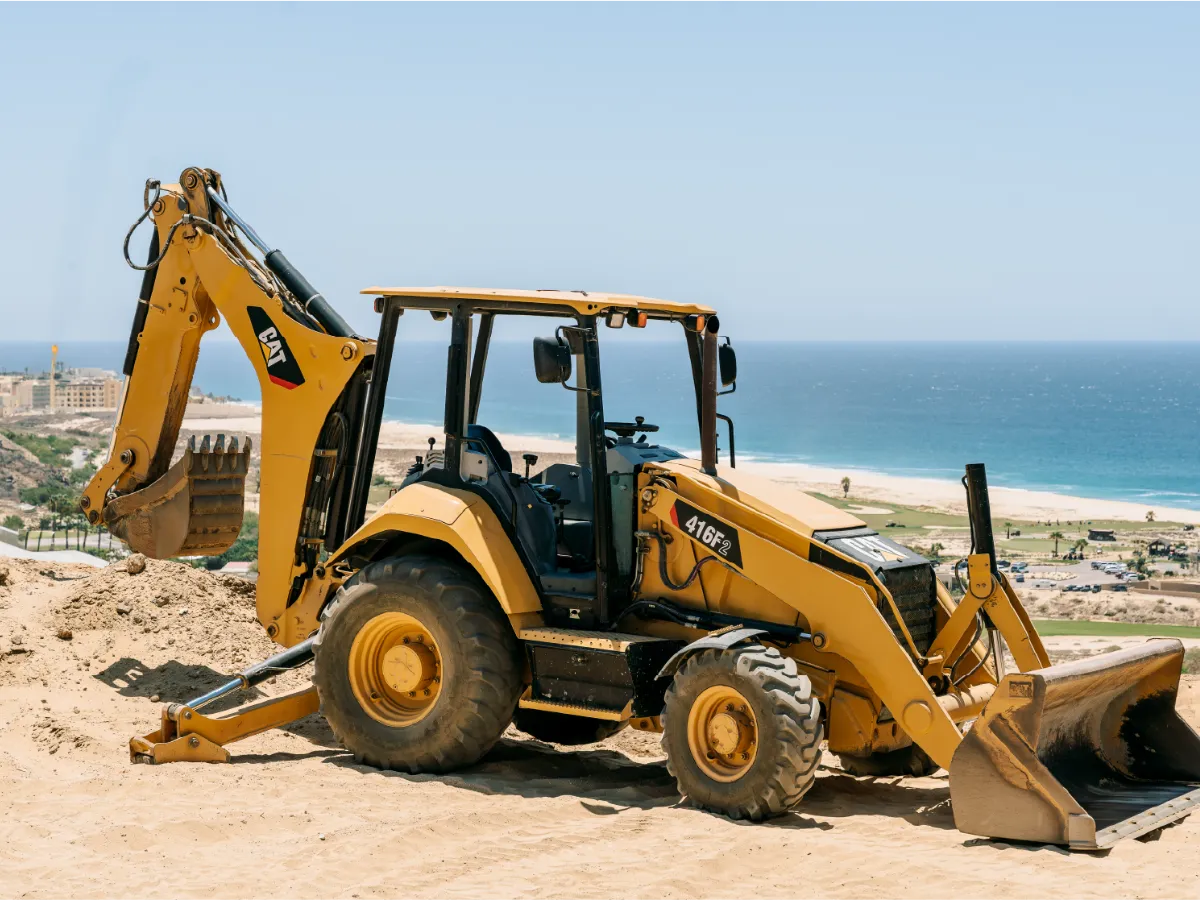 CAT 416F2 backhoe loader parked on sand, with the beach and coastline behind it